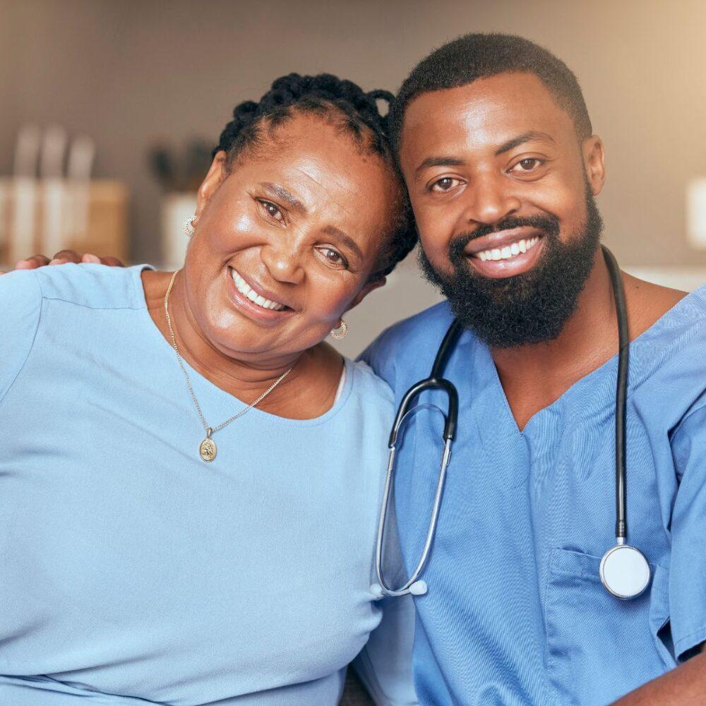 Black senior woman sitting comfortably with Black doctor or nurse wearing a stethoscope in what appears to be a home environment.