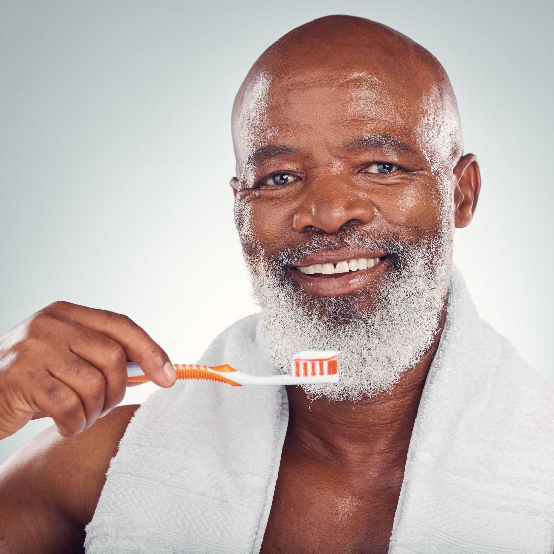 An attractive Black senior man with a beard smiling while holding a toothbrush with toothpaste.