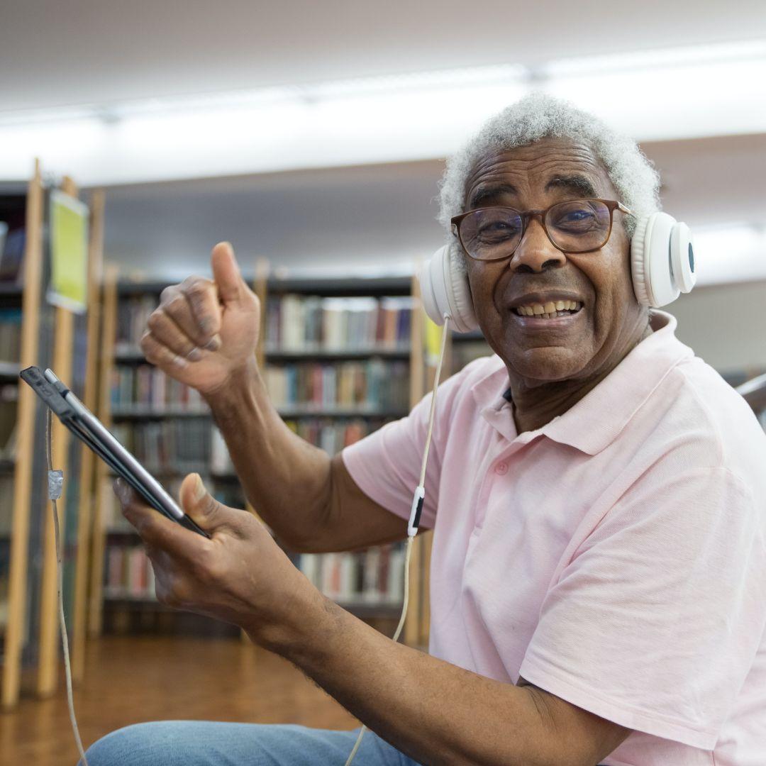 A smiling Black man in the local library listening to something on a tablet while wearing headphones.