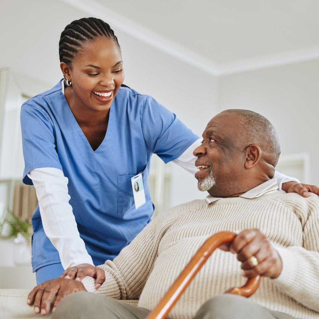 A Black senior man holding a cane while sitting on a comfortable sofa and smiling at a nurse who has her arm on his shoulder.