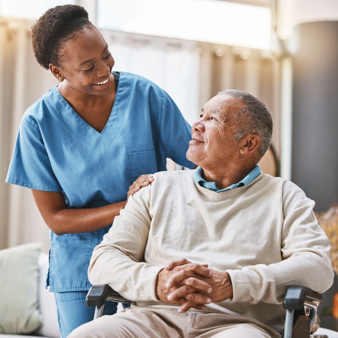 A smiling Black senior man in a wheelchair with a young Black nurse in what appears to be a nursing home environment.