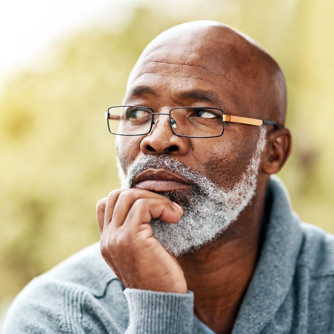 A handsome senior Black man wearing glasses and sitting outdoors with his hand on his chin as if he is pondering something.