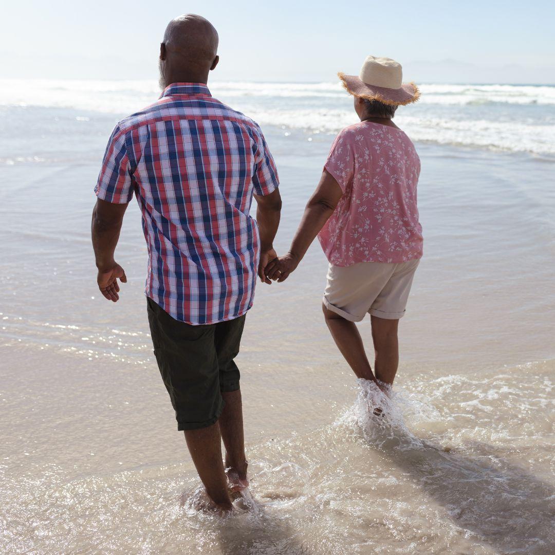 A senior Black couple holding hands and walking on a pretty beach on a sunny day.