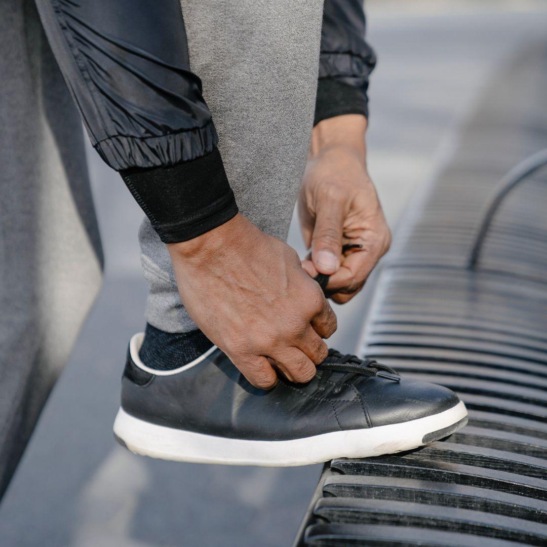 A Black senior man tying his shoe on a park bench while wearing sweatpants and a jacket.