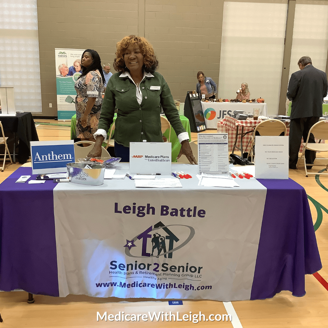 Photo of Leigh Battle, Medicare Advisor and Educator, standing behind a branded Senior 2 Senior table at a vendor fair.